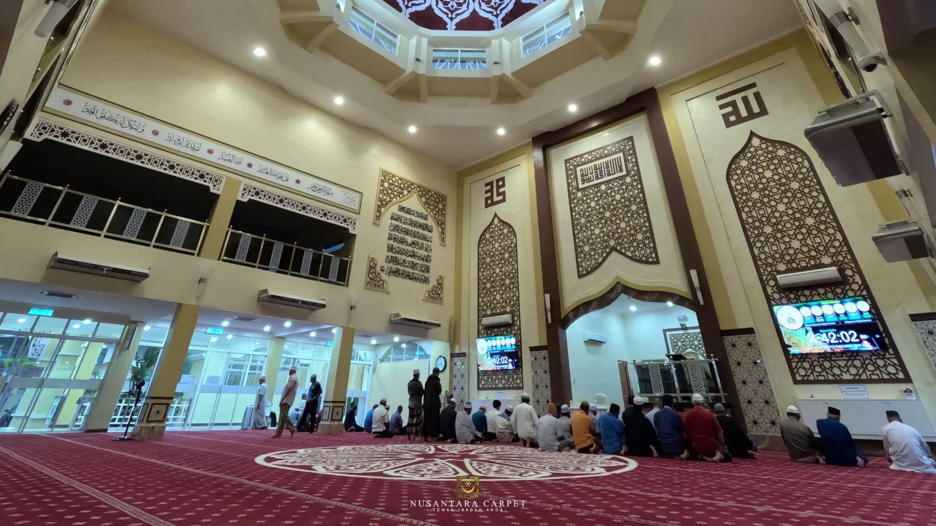 Pemandangan sudut pandang luas memaparkan ruang dewan solat dua tingkat yang dipenuhi jemaah berlantai hamparan hasil siap pemasangan karpet masjid gred premium berwarna merah marun merah dengan corak garisan saf lurus bertemakan motif geometri islamik kubah di Masjid Sentosa Teras Jernang, Bangi, Selangor, menunjukkan kawasan mihrab berbingkai panel kayu coklat besar di hadapan, jam digital waktu solat elektronik di dinding kanan, serta struktur kubah tengah siling yang tinggi berhiaskan seni khat ayat suci.