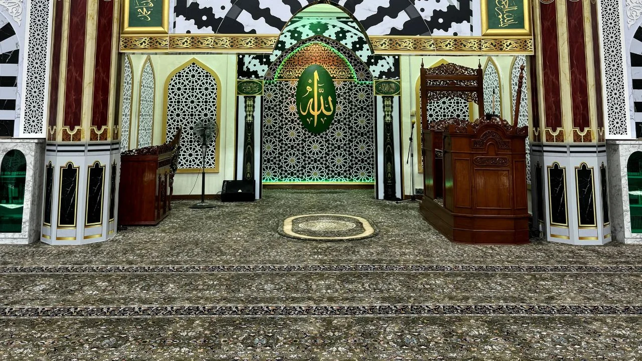 Pemandangan ruang hadapan mihrab memaparkan hasil siap pemasangan karpet surau custom-made gred premium berwarna dasar kelabu gelap dengan corak abstrak geometri kerawang emas dan barisan saf lurus di Surau Khalid Al-Walid IPK Selangor, Shah Alam, menunjukkan mimbar kayu coklat gelap tradisional di sebelah kanan, lambang kaligrafi ayat suci Allah hijau keemasan di tengah dinding mihrab berhias ukiran batu putih, serta struktur tiang marmar besar di bahagian kiri dan kanan.