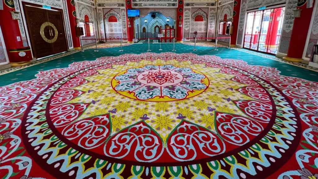 Pemandangan sudut luas (wide-angle view) memaparkan hasil siap pemasangan karpet masjid kustom gred premium berwarna dasar hijau tua di bahagian ruang solat utama Masjid Al-Badr (Masjid Cina), memberikan fokus kepada reka bentuk motif bulatan medallion mega berskala besar di bahagian tengah yang dipenuhi lingkaran geometri Islamik serta corak awan tradisional gabungan warna merah marun, kuning keemasan, merah jambu, dan putih, bersanding dengan struktur hiasan dinding mihrab bertemakan seni bina Cina Muslim merah putih emas di bahagian belakang, pintu gerbang kayu coklat di sebelah kiri, serta tingkap cermin berbingkai merah di sebelah kanan