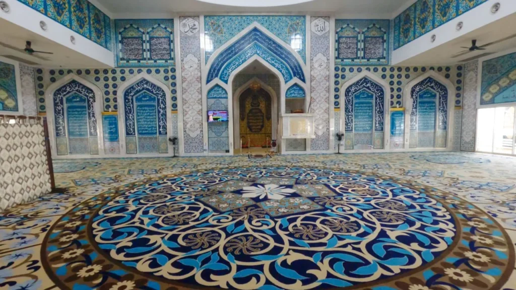 Pemandangan sudut luas dari aras lantai memaparkan keindahan ruang dalaman dewan solat utama berlantai hamparan karpet masjid kustom gred premium rona coklat keemasan, mempamerkan reka bentuk corak motif bulatan medallion mega berskala besar berwarna biru diraja dan biru toska di bahagian tengah yang dipenuhi lingkaran geometri rumit berpusatkan bunga bintang putih, bersanding harmoni dengan hiasan dinding mihrab serta panel pintu gerbang melengkung tajam di bahagian belakang yang sarat dengan seni bina jubin seramik kaligrafi al-Quran bertema rona biru putih keemasan, berserta papan skrin televisyen kecil di tiang konkrit petak putih dan deretan kipas siling hitam di bahagian atas kiri kanan