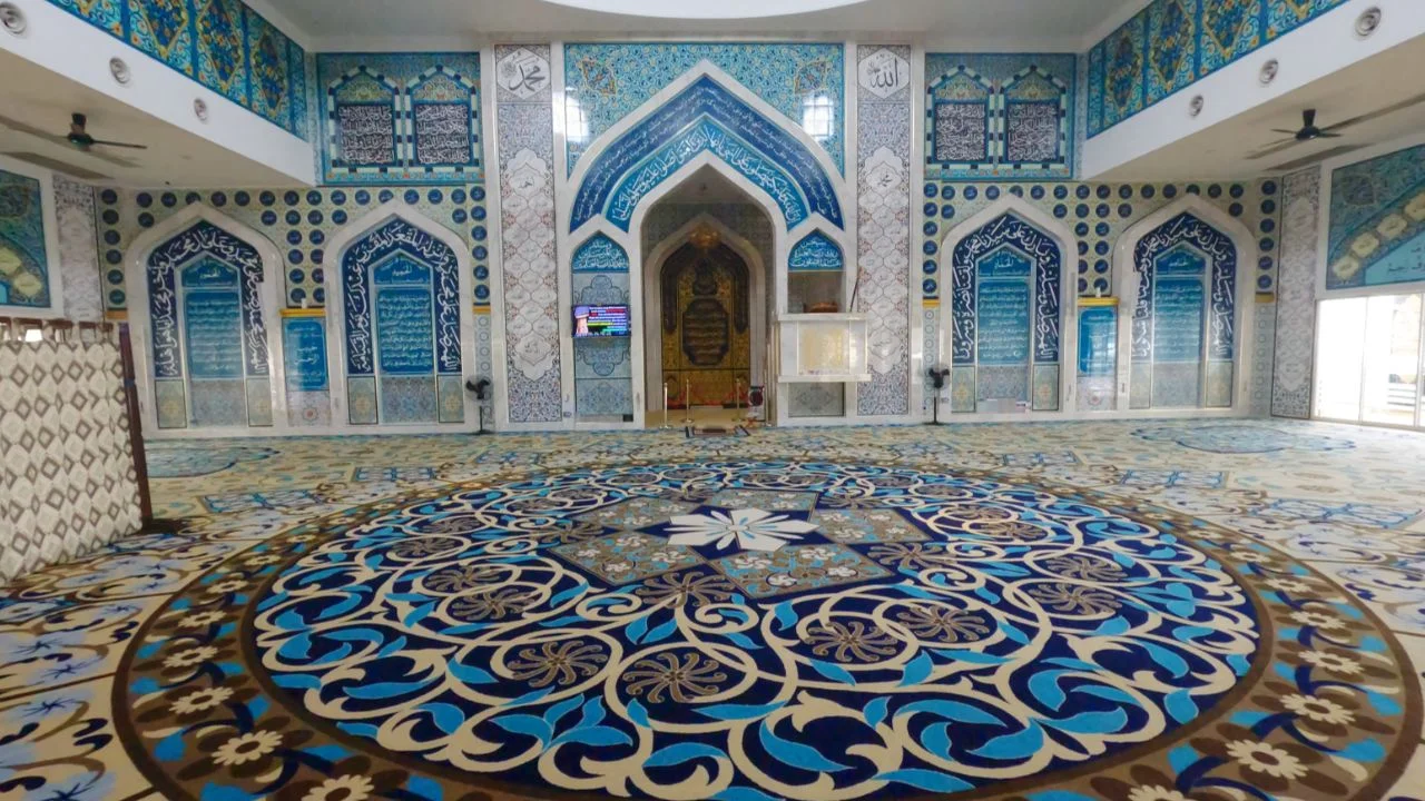 Pemandangan sudut luas dari aras lantai memaparkan keindahan ruang dalaman dewan solat utama berlantai hamparan karpet masjid kustom gred premium rona coklat keemasan, mempamerkan reka bentuk corak motif bulatan medallion mega berskala besar berwarna biru diraja dan biru toska di bahagian tengah yang dipenuhi lingkaran geometri rumit berpusatkan bunga bintang putih, bersanding harmoni dengan hiasan dinding mihrab serta panel pintu gerbang melengkung tajam di bahagian belakang yang sarat dengan seni bina jubin seramik kaligrafi al-Quran bertema rona biru putih keemasan, berserta papan skrin televisyen kecil di tiang konkrit petak putih dan deretan kipas siling hitam di bahagian atas kiri kanan