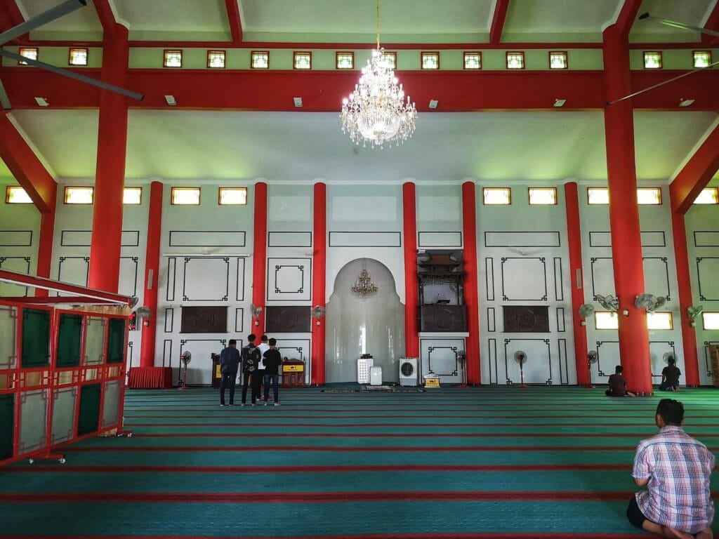 Pandangan luas ruang solat utama memaparkan hamparan karpet masjid tebal berwarna merah marun dengan garisan saf kuning bercorak kustom bulat geometri emas, dikelilingi oleh dinding panel kayu coklat gelap terukir tulisan jawi kaligrafi Arab, struktur replika mimbar bumbung cina pagoda kayu, serta siling berkotak hiasan geometri bercahaya lampu dinding suam