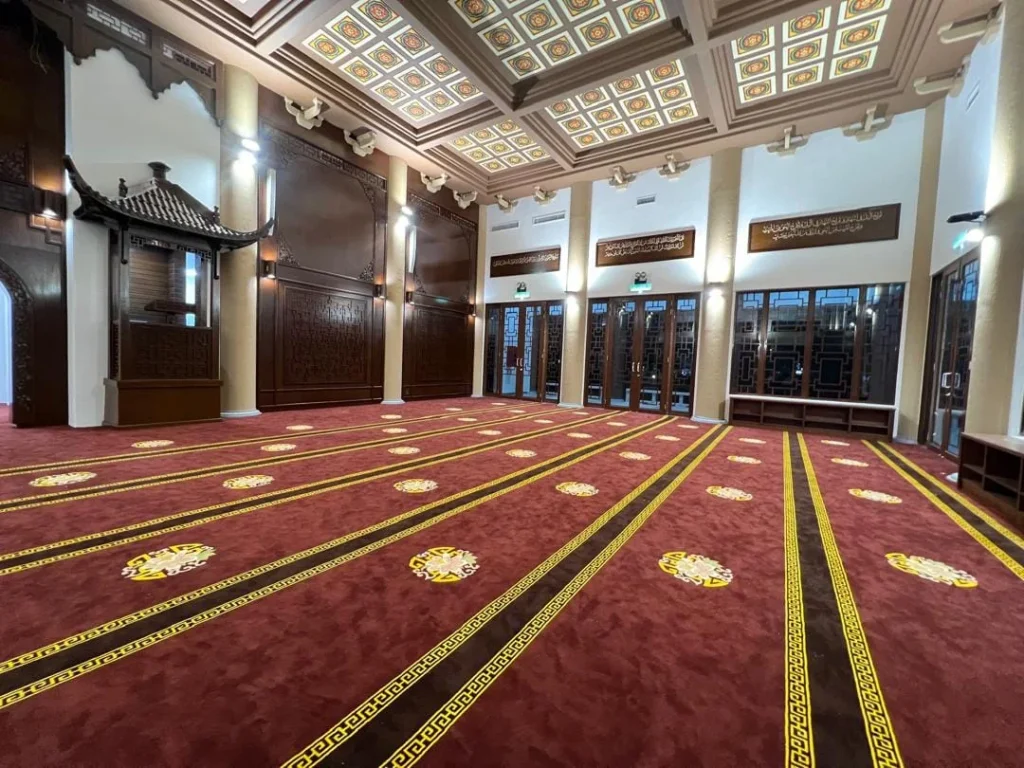 Pandangan luas ruang solat utama memaparkan hamparan karpet masjid tebal berwarna merah marun dengan garisan saf kuning bercorak kustom bulat geometri emas, dikelilingi oleh dinding panel kayu coklat gelap terukir tulisan jawi kaligrafi Arab, struktur replika mimbar bumbung cina pagoda kayu, serta siling berkotak hiasan geometri bercahaya lampu dinding suam