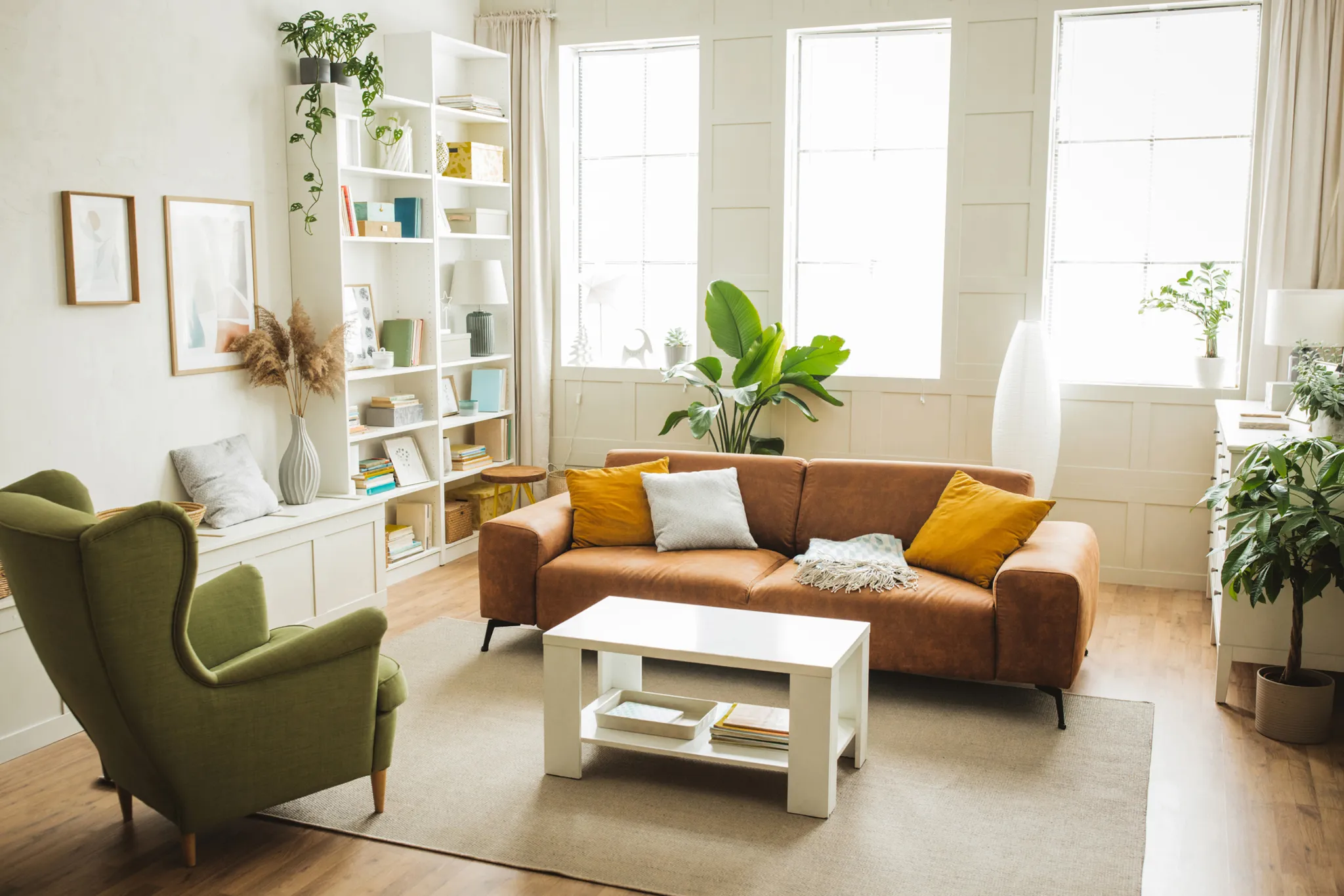 Ruang tamu pencahayaan terang dari tiga tingkap memaparkan sebuah sofa panjang kain warna coklat dengan bantal hiasan kuning dan kelabu, sebuah kerusi lengan santai warna hijau di bahagian tepi, sebuah meja kopi putih rendah di atas hamparan karpet polos warna krim, rak buku putih tinggi bersusun barangan hiasan, serta pasu pokok hijau di lantai kayu