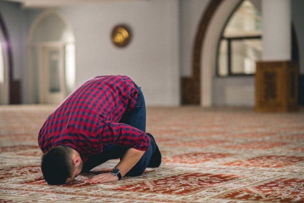 Pandangan jarak dekat dari sudut tepi serong memaparkan seorang lelaki bermabut pendek memakai kemeja petak-petak warna merah hitam dan seluar biru tua sedang bersujud menunaikan solat di atas hamparan karpet masjid tebal berwarna merah marun bercorak putih emas, berlatarbelakangkan ruang dalaman surau yang luas beralaskan tiang konkrit putih dan pintu gerbang melengkung kayu