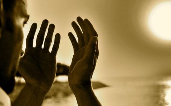 Pandangan jarak dekat siluet separuh muka seorang lelaki dari arah tepi sedang menadah kedua-dua belah tangan untuk berdoa menghadap pancaran cahaya matahari yang terang dengan latar belakang suasana senja keemasan pudar