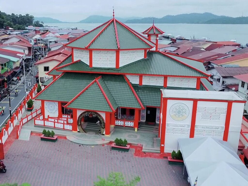 Pemandangan dari atas (aerial view) kompleks bangunan Masjid Al-Khairiyah di Pulau Pangkor Perak yang menampilkan reka bentuk seni bina pagoda tradisional Cina dengan struktur bumbung bertingkat warna hijau, dinding berwarna merah dan putih, pintu gerbang membulat hitam emas di bahagian hadapan, dataran pejalan kaki berturap bata merah, struktur bangunan putih di bahagian kanan berserta khemah putih, berlatarbelakangkan kawasan petempatan rumah perkampungan nelayan dan pemandangan laut serta bukit hijau di bahagian belakang bawah langit mendung