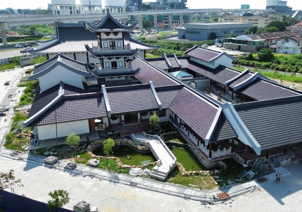 Pemandangan dari atas (aerial view) kompleks bangunan Masjid Jamek Cina Muslim di Klang yang menampilkan reka bentuk seni bina tradisional Cina dengan bumbung bertingkat warna kelabu gelap, struktur menara pagoda di tengah, jambatan konkrit putih melintasi kolam air kecil di bahagian hadapan, serta kawasan persekitaran jalan raya dan landasan kereta api di bahagian belakang bawah sinaran cahaya matahari