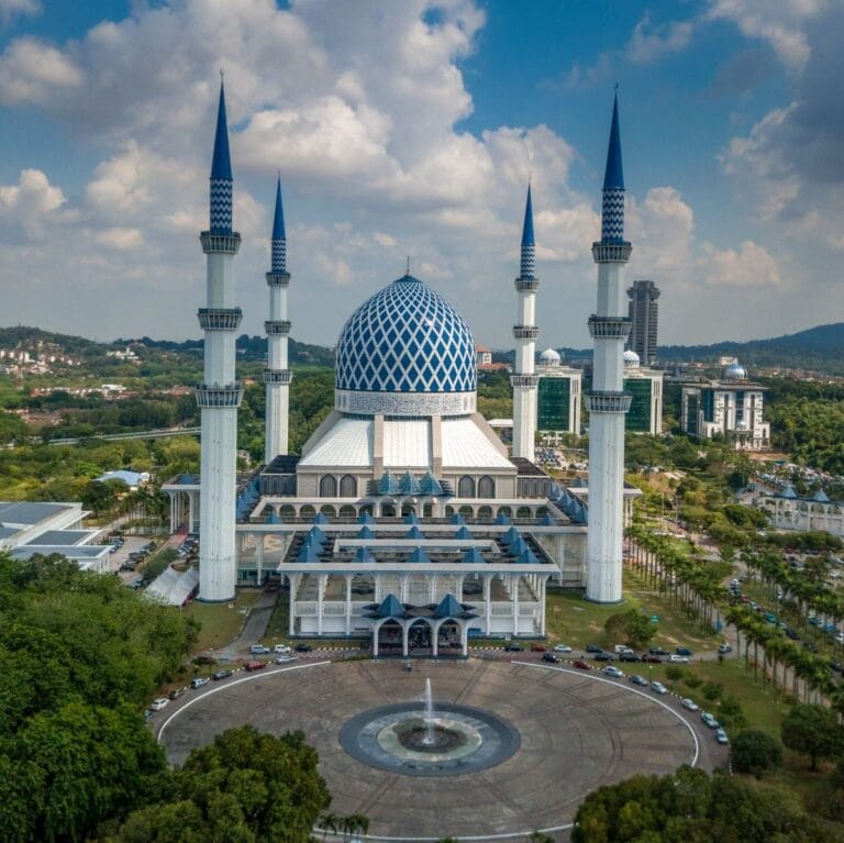 Pemandangan dari atas (aerial view) struktur penuh bangunan Masjid Sultan Salahuddin Abdul Aziz Shah di Shah Alam yang memaparkan sebuah kubah besar bercorak geometri biru putih, empat buah menara tinggi berwarna putih dengan hujung biru, kawasan dataran bulat berpusat air pancut di bahagian hadapan, serta landskap pokok hijau di bawah langit berawan