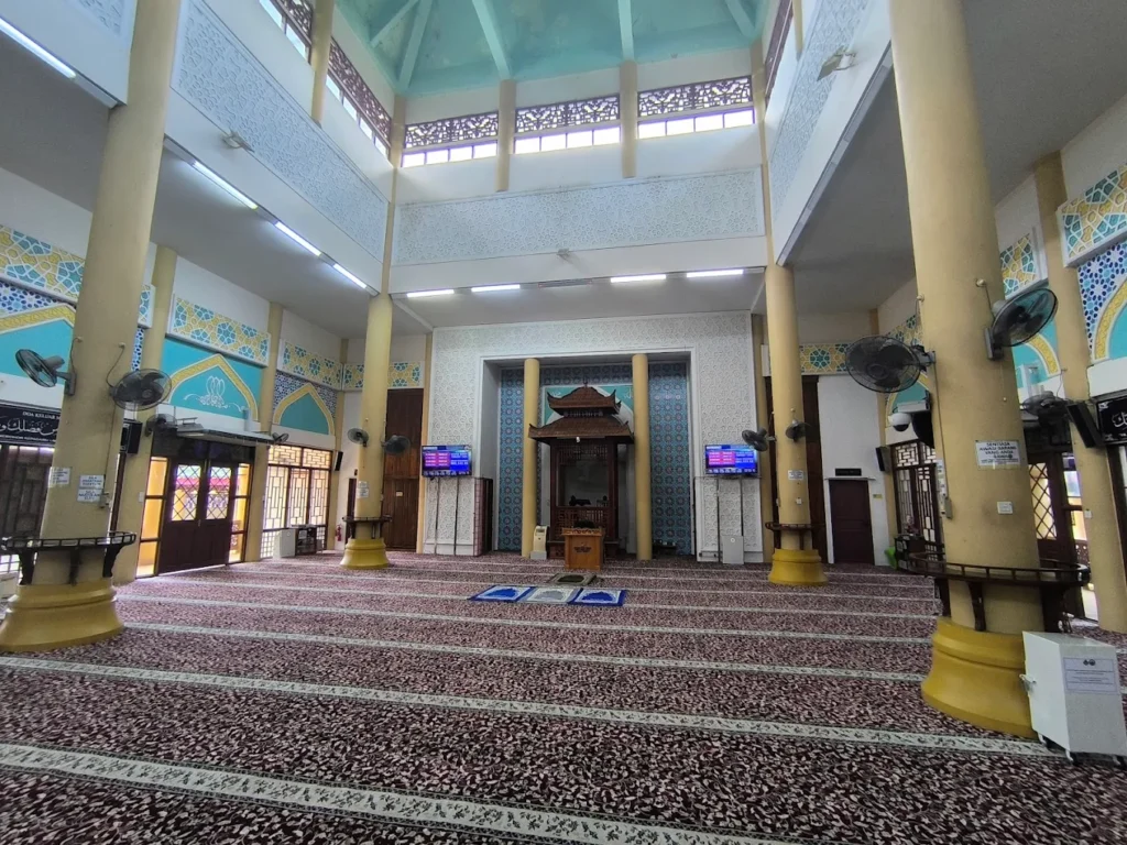 Pandangan luas ruang solat utama Masjid Jubli Perak Sultan Ismail Petra di Rantau Panjang Kelantan memaparkan hamparan karpet saf tebal berwarna ungu bercorak geometri putih emas, disokong oleh tiang-tiang silinder besar berwarna kuning krim, struktur mihrab putih dengan hiasan corak dinding biru, sebuah mimbar kayu coklat gelap berbumbung melengkung gaya pagoda di tengah mihrab, skrin paparan digital TV di kiri kanan, rekaan gerbang pintu kayu berbingkai kaca berwarna-warni di bahagian kiri, serta hiasan corak geometri Islamik biru kuning di dinding bawah siling berkubah tinggi