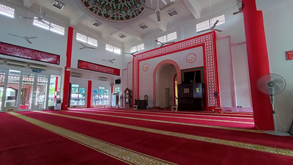 Pandangan luas dari sudut tepi memaparkan ruang solat utama Masjid Cina Negeri Melaka di Krubong yang dihampar permaidani saf berwarna merah dengan jalur garisan kuning keemasan, disokong oleh tiang bulat besar berwarna merah silinder, struktur gerbang mihrab melengkung berbingkai corak geometri merah putih, sebuah mimbar kayu coklat gelap di dalam mihrab, jam berdiri antik kayu, deretan kipas syiling putih, serta pintu cermin kaca berbingkai putih di bahagian kiri bawah siling putih bercorak bulat geometri