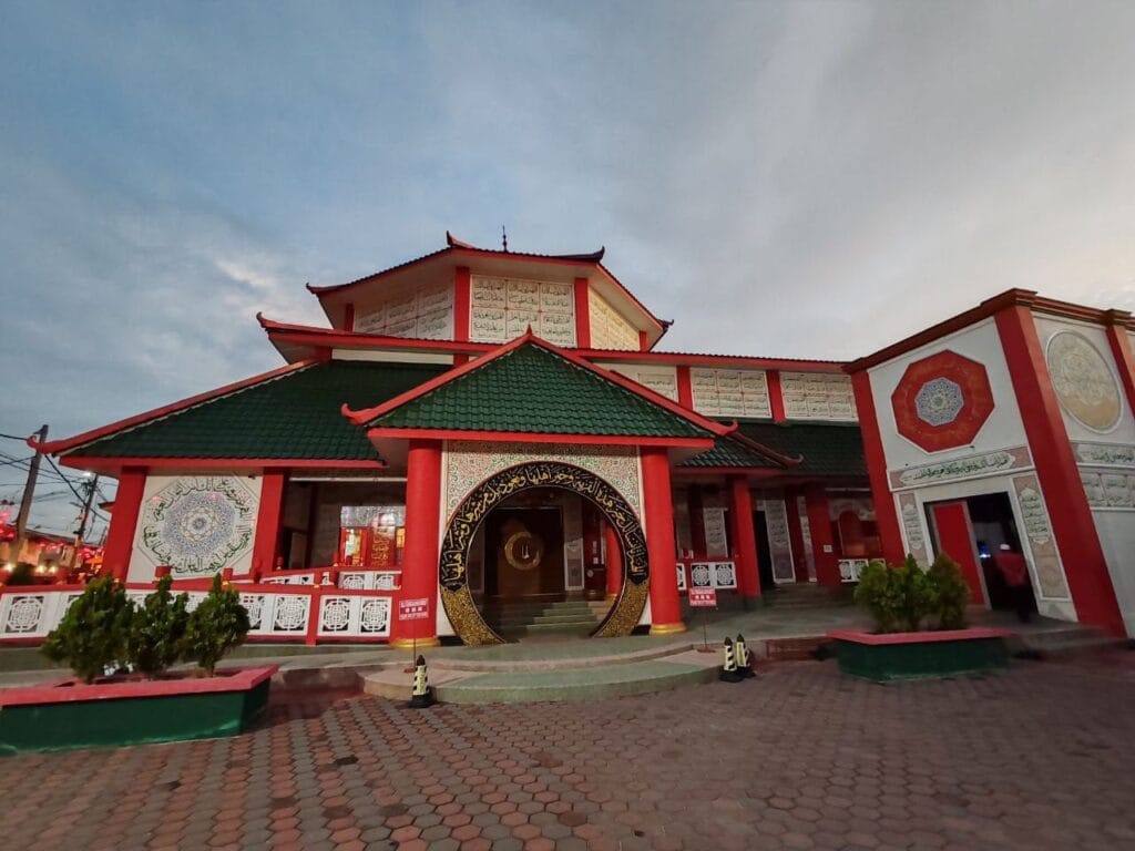 Pemandangan luar hadapan kompleks bangunan Masjid Al-Khairiyah di Segamat Johor yang memaparkan reka bentuk seni bina pagoda tradisional Cina dengan struktur gerbang bulat pintu masuk besar berwarna hitam emas terukir ayat suci Al-Quran tulisan khat Arab, dua tiang silinder merah di hadapan, dinding berwarna merah dan putih, bumbung berlapis warna hijau gelap dengan rekaan bucu melengkung merah, papan panel putih terukir teks jawi di bahagian atas, pagar konkrit putih bercorak tradisional di bahagian tepi, serta pokok renek hiasan di dalam pasu bata berlatarbelakangkan lantai pejalan kaki berturap bata heksagon merah di bawah langit petang yang cerah