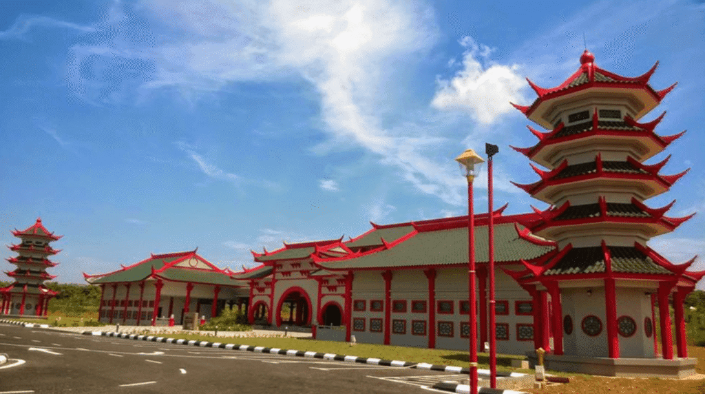 Pemandangan luar kompleks bangunan Masjid Cina Negeri Melaka di Krubong yang memaparkan seni bina tradisional Cina dengan dinding putih berbingkai merah, bumbung berlapis warna hijau gelap dengan hujung melengkung merah, struktur menara pagoda bertingkat di bahagian hadapan, tiang lampu jalan berwarna merah emas, serta laluan jalan raya berturap dengan garisan pembahagi hitam putih di bawah langit biru berawan