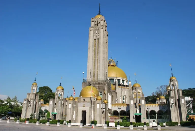 Pemandangan luar Masjid DiRaja Sultan Suleiman di Klang memaparkan struktur bangunan konkrit kelabu dengan menara tinggi di tengah, kubah-kubah berwarna kuning keemasan, dan deretan tiang pagar di bahagian hadapan di bawah langit biru yang cerah