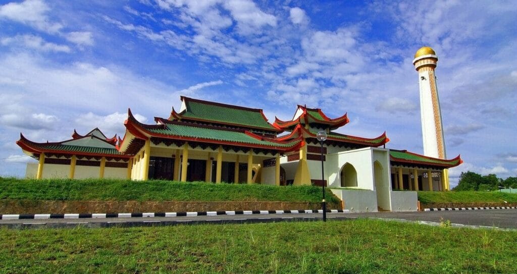 Pemandangan luar kompleks bangunan Masjid Jubli Perak Sultan Ismail Petra di Rantau Panjang Kelantan yang memaparkan seni bina tradisional Cina dengan tiang-tiang bulat kuning, bumbung berlapis warna hijau dengan bucu melengkung merah, sebuah menara tinggi berwarna putih dengan kubah kecil berwarna emas di bahagian kanan, kawasan benteng berumput hijau di hadapan, serta laluan jalan raya berturap dengan pembahagi jalan hitam putih di bawah langit biru cerah berawan putih