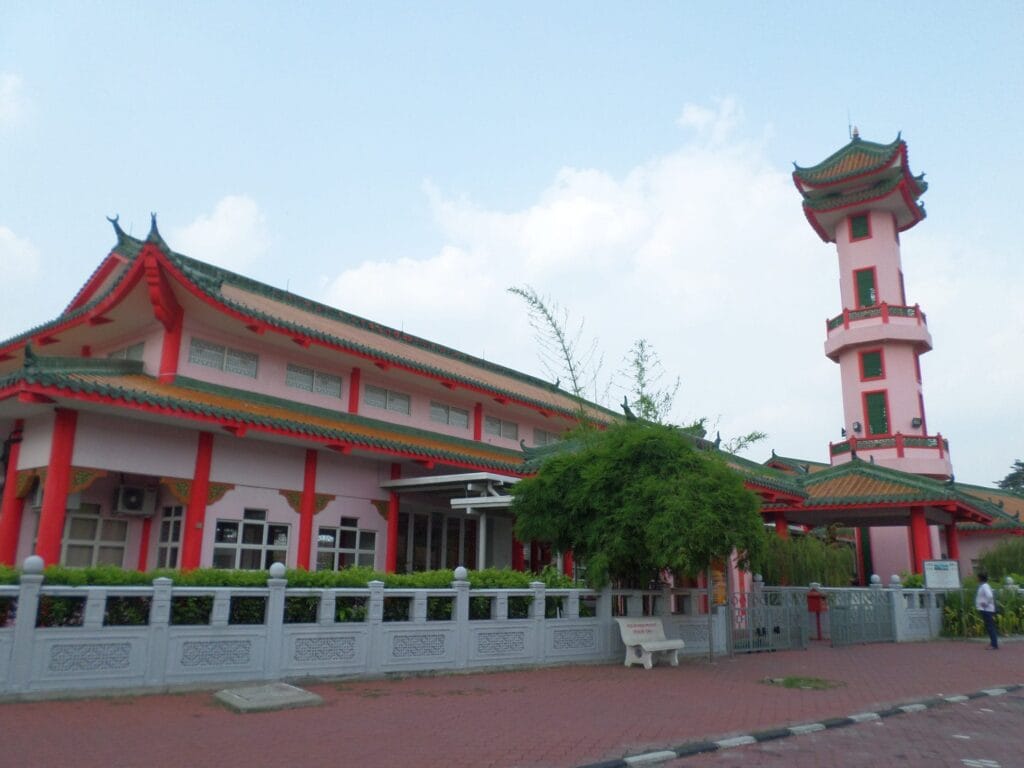 Pemandangan luar kompleks bangunan Masjid Muhammadiah di Ipoh Perak yang memaparkan seni bina tradisional Cina dengan dinding berwarna merah jambu pudar, struktur tiang merah, bumbung berlapis warna hijau dengan bucu melengkung merah, sebuah menara pagoda tinggi bercorak putih merah jambu di bahagian kanan, pagar konkrit kelabu berukir motif tradisional di sepanjang hadapan, sebatang pokok hijau rimbun di tepi bangku putih, serta seorang individu berjalan di bahagian tepi bawah langit cerah berawan pudar