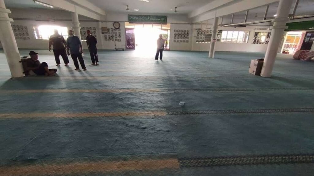 Ruang dalaman Masjid At-Taqwa yang luas beralaskan karpet lama berjalur hijau pudar tersusun saf dengan kelihatan beberapa orang jemaah lelaki sedang berdiri dan duduk bersandar pada tiang bangunan