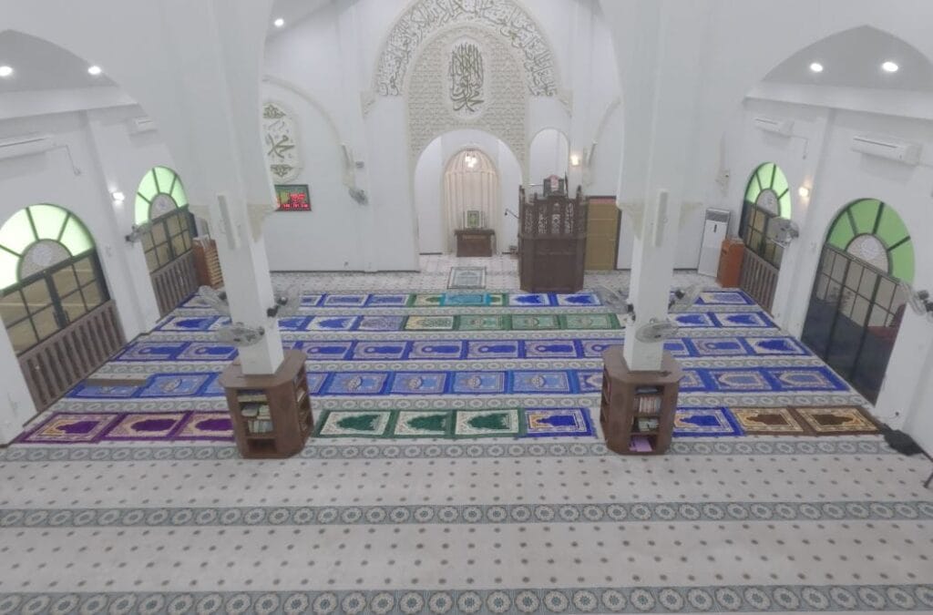Pandangan dari sudut tinggi dewan solat Masjid Jamek Matang Gerdu memaparkan kombinasi sejadah saf individu berwarna-warni dan karpet saf hijau lembut