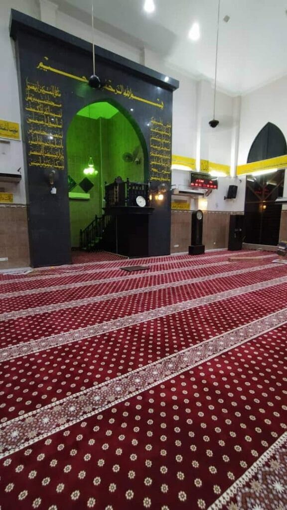 Pandangan pepenjuru ruang solat Masjid Jamek Bagan Dalam memaparkan struktur mihrab hitam berkubah hijau dan hamparan karpet saf merah hati