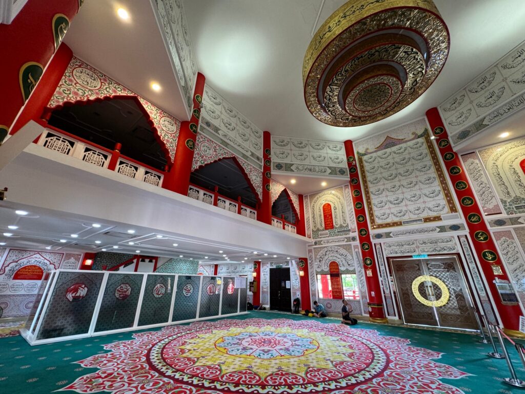 Pandangan luas dewan solat Masjid Al-Khairiah memaparkan hiasan kaligrafi khat, tiang merah megah, dan hamparan karpet kustom bercorak medallion besar yang meriah