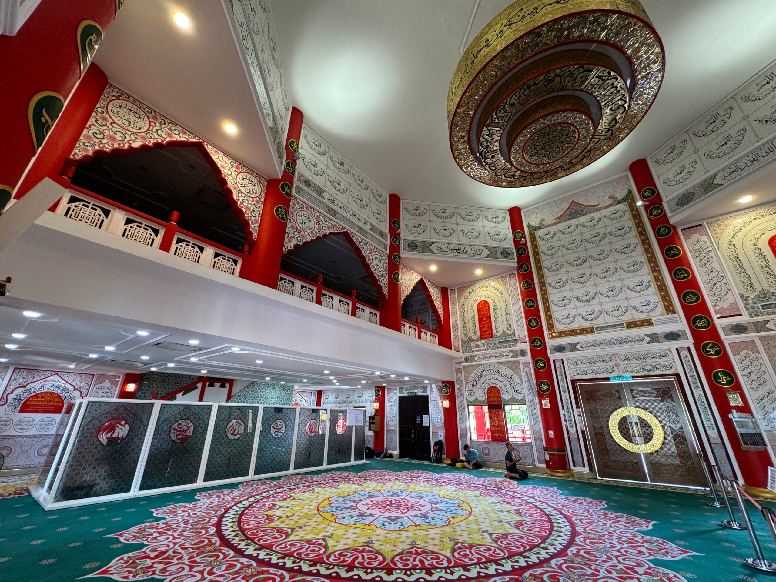 Pandangan luas dewan solat Masjid Al-Khairiah memaparkan hiasan kaligrafi khat, tiang merah megah, dan hamparan karpet kustom bercorak medallion besar yang meriah