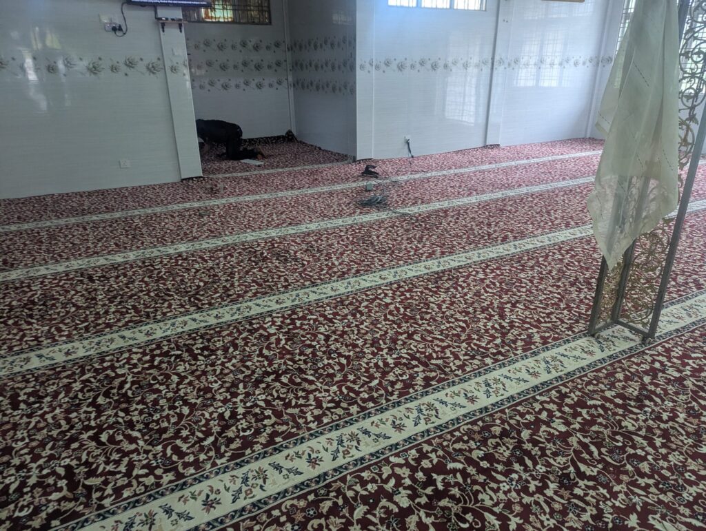 Karpet merah bercorak bunga dengan jalur saf krim sedang dipasang di ruang solat Surau An Nur Pandan Mewah