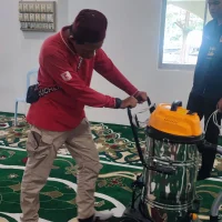 Gambar foto dokumentasi memaparkan seorang lelaki daripada pasukan teknikal Nusantara Carpet berkopiah merah hati, berbaju kemeja lengan panjang merah dengan beg silang hitam bertulis KUCHEN, serta berseluar kargo coklat muda sedang membongkok memegang pemegang besi untuk mengendalikan jentera vakum industri silinder keluli besar berkepala jingga kuning bertayar roda, di atas permukaan hamparan karpet masjid hijau tebal premium bercorak geometri bunga putih kecil saf lurus bersanding dengan rekaan motif daun merah putih besar yang menjalar di bahagian bawah, manakala di sebelah kanan belakang kelihatan kelibat seorang lelaki lain berbaju hitam sedang berdiri memerhatikan proses ujian tersebut di dalam bilik ruang solat berhampiran sebuah jam dinding bulat petunjuk pukul dua suku