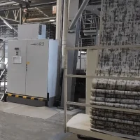 Pemandangan jarak dekat di dalam dewan pembuatan industri memaparkan sebuah kabinet panel kawalan elektrik besar berwarna kelabu putih bertulis kod CRP3 berkaki asas hitam kuning yang dilengkapi suis kawalan keselamatan di bahagian kiri pekarangan lantai konkrit kelabu, manakala di bahagian kanan hadapan menunjukkan struktur troli pemuat besi putih petak yang sarat dengan tindihan lapisan lipatan fabrik karpet masjid bercorak abstrak geometri kombinasi warna hitam, kelabu, dan putih pudar yang sedang disalurkan turun secara menegak dari tingkat atas jentera penenunan komputer