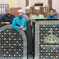 Gambar foto dokumentasi memaparkan barisan sembilan orang lelaki Muslim berdiri bersebelahan memakai baju melayu, jubah, dan kopiah di dalam ruang solat berlantai jubin corak Islamik biru putih dengan mimbar kayu dan jam berdiri besar di bahagian belakang, menunjukkan ahli jawatankuasa kariah memegang dan memamerkan dua unit sejadah kustom eksklusif berlatarkan warna biru tua corak geometri bintang emas, dengan sejadah di sebelah kiri berbentuk melengkung kubah manakala sejadah di sebelah kanan memaparkan tenunan khas grafik kubah hijau putih serta teks tulisan rumi putih berbunyi Masjid Jamek Bandar Kulai