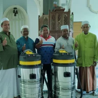 Gambar foto dokumentasi berlatarbelakangkan ruang solat memaparkan barisan tujuh orang lelaki berdiri bersebelahan di atas hamparan karpet masjid bercorak hijau putih pudar dengan mimbar kayu coklat tua di bahagian belakang, menunjukkan dua orang wakil teknikal Nusantara Carpet memakai jaket jingga hitam dan kemeja korporat kelabu kuning bersama lima orang ahli kariah jawatankuasa masjid berbaju melayu serta berkain pelikat yang kesemuanya sedang menunjukkan isyarat tanda bagus ibu jari di hadapan dua unit mesin vakum industri silinder keluli besar berkepala kuning hitam