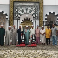 Gambar foto dokumentasi jarak dekat memaparkan barisan sepuluh orang lelaki Muslim termasuk kru Nusantara Carpet dan ahli jawatankuasa kariah berdiri tegak sebaris di atas hamparan karpet masjid tebal premium bercorak geometri bunga kelabu hijau lumut dengan rekaan motif medallion bulat keemasan besar yang mewah di bahagian hadapan bawah, menunjukkan sebahagian mereka memakai baju melayu, jubah, kopiah putih, bersongkok, dan pakaian kasual, bersanding bersama dua unit mesin vakum industri pembersih warna merah hitam keluli tahan karat di tengah-tengah mereka berlatarbelakangkan struktur dinding mihrab yang megah bercat kelabu putih dengan ukiran hiasan geometri Islamik serta plak kaligrafi al-Quran hitam keemasan melengkung tajam tepat di bahagian atas tengah