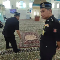 Gambar foto dokumentasi memaparkan suasana di dalam ruang solat utama berlantai karpet masjid hijau tebal bermotif corak geometri bunga tradisional bersambung saf lurus, menunjukkan seorang pegawai kanan polis berpakaian seragam operasi hitam lengkap bersongkok rasmi dengan pingat perkhidmatan sedang berdiri menghadap ke arah kanan di bahagian depan, manakala seorang lagi pegawai polis berbaju hitam sedang membongkok meninjau hamparan sejadah kustom kecil bermotif medallion bulat coklat keemasan di tengah lantai saf, serta kelihatan beberapa orang jemaah lelaki Muslim sedang duduk bersila dan berdiri di kawasan belakang berhampiran mimbar kayu coklat tua