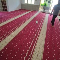 Gambar foto dokumentasi sudut pandangan luas dari dalam bilik memaparkan hasil kerja pemasangan karpet surau berwarna merah menyala dengan corak saf lurus berjalur putih pudar keemasan bermotif bunga geometri Islamik tradisional, menunjukkan susunan garisan saf yang selari merentangi seluruh ruang lantai dari pekarangan pintu gelongsor kaca di sebelah kanan hingga ke bahagian dinding hadapan bercat putih, kelihatan kelibat seorang lelaki Muslim berseluar hitam dan berbaju t-shirt kelabu sedang berjalan berkaki ayam di atas saf bahagian kanan bawah naungan pencahayaan lampu siling serta kipas berpusing