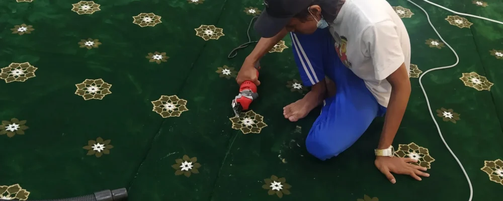 Seorang pekerja krew teknikal Nusantara Carpet memakai topi hitam dan baju putih sedang duduk melutut melakukan proses ukiran kemasan akhir menggunakan mesin pemotong elektrik khas di atas permukaan karpet masjid custom-made gred premium berwarna hijau tua bercorak bunga geometri bintang emas dan barisan saf lurus, menunjukkan urutan wayar putih yang merintangi lantai dewan solat berlatarbelakangkan dinding putih.