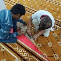 Pemandangan sudut atas (top-down view) memaparkan dua orang pekerja lelaki daripada pasukan teknikal Nusantara Carpet sedang melutut di atas hamparan karpet masjid tebal berwarna coklat keemasan bermotif corak geometri bunga saf lurus bertanda corak putih emas di bahagian tepi, menunjukkan pekerja di sebelah kiri memakai baju sweater sejuk rajutan berjalur biru putih manakala pekerja di sebelah kanan memakai kemeja t-shirt putih berseluar cerah, kedua-duanya sedang fokus bekerjasama menggunakan alat pemotong besi manual untuk memotong bahagian hujung bucu karpet mengikut acuan bentuk tiang konkrit petak putih pekarangan masjid bagi menampakkan bahagian pelapik underlay foam berwarna merah jambu cerah di bawahnya
