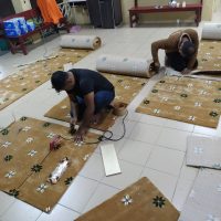 Gambar foto dokumentasi dari sudut pandangan atas memaparkan dua orang pekerja lelaki daripada pasukan pemasangan Nusantara Carpet sedang bertelut di atas lantai jubin kelabu muda untuk menguruskan hamparan kepingan gulungan karpet surau tebal berwarna coklat keemasan bermotif corak geometri bunga bintang putih hitam, menunjukkan pekerja di sebelah kiri memakai baju t-shirt hitam berseluar jeans biru sedang menggunakan alat seterika besi pemanas haba untuk mencairkan pita pelekat penyambung gam khas di bawah garisan saf, manakala pekerja di sebelah kanan memakai kemeja lengan panjang coklat sedang membongkok merapatkan bucu potongan fabrik kain, berlatarbelakangkan suasana dalaman bilik surau yang dilengkapi deretan bangku kayu coklat merah serta papan kenyataan dinding di bahagian belakang