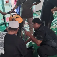 Gambar foto dari sudut pandangan atas memaparkan suasana di dalam ruang solat berlantai karpet masjid hijau tebal bermotif corak bunga geometri putih merah, menunjukkan seorang wakil teknikal Nusantara Carpet berbaju hitam sedang bertelut memberikan tunjuk ajar cara mengendalikan mesin vakum industri silinder keluli berkepala jingga hitam kepada seorang ahli jawatankuasa masjid berkopiah putih yang duduk bersila di hadapannya, sambil disaksikan oleh tiga orang lelaki lain yang berdiri memerhati di pekarangan pintu cermin kaca