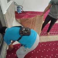 Gambar foto dokumentasi sudut pandangan atas memaparkan dua orang pekerja lelaki daripada pasukan teknikal Nusantara Carpet sedang menguruskan pemasangan karpet surau merah bercorak saf lurus geometri putih di dalam bilik, menunjukkan seorang pekerja berbaju t-shirt biru cerah memakai beg silang hitam sedang membongkok melutut di bahagian bawah kiri menggunakan pisau utiliti untuk memotong bucu permaidani mengikut acuan bingkai pintu kaca, manakala seorang lagi pekerja berdiri di bahagian atas kanan memakai baju kelabu kemeja t-shirt berseluar hitam sedang memegang gulungan karpet contoh berasingan berwarna coklat muda bercorak saf berhampiran sebuah mesin vakum industri pembersih kecil berwarna biru putih yang diletakkan di tepi dinding konkrit putih